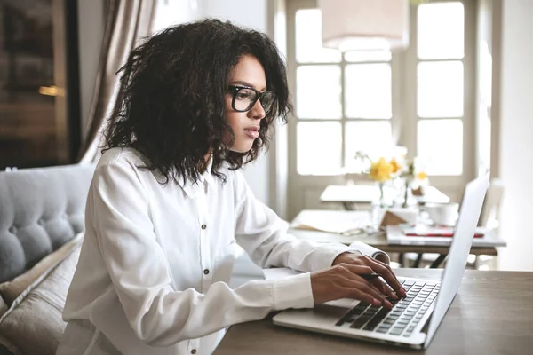 Hermosa señora que trabaja en el ordenador portátil en restaurante.Pretty chica afroamericana en gafas de escribir en el ordenador en la cafetería —  Fotos de Stock