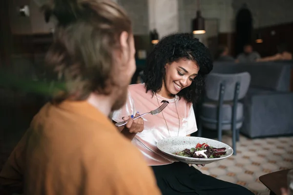 Chica sonriente sentada en el restaurante con un amigo. Bonita dama afroamericana sentada en la cafetería con plato de ensalada en la mano — Foto de Stock