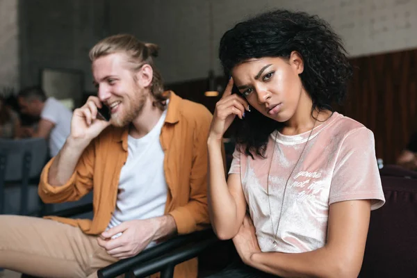 Porträt eines jungen Mannes und eines jungen Mädchens im Restaurant. aufgebrachte afrikanisch-amerikanische Mädchen mit dunkelgelockten Haaren traurig in die Kamera schauen, während Junge mit blonden Haaren fröhlich auf seinem Handy im Café reden — Stockfoto