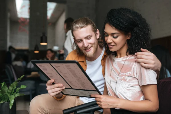 Niza chica afroamericana con pelo rizado oscuro y chico con el pelo rubio felizmente buscando en el menú en la cafetería —  Fotos de Stock