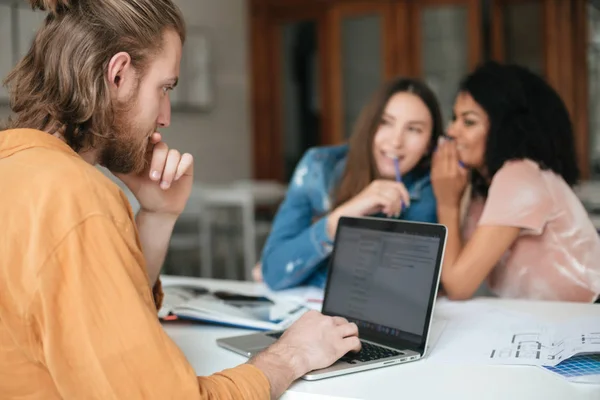 Retrato de jovem com cabelo loiro e barba sentado no escritório e trabalhando em seu laptop, enquanto duas meninas fofocando no fundo — Fotografia de Stock