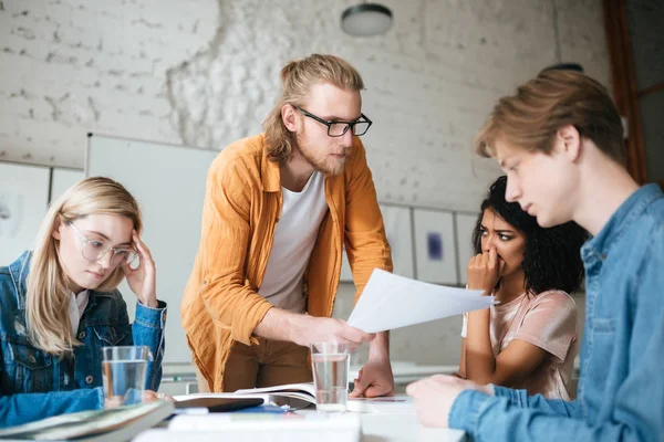 Ritratto di giovane insegnante serio in occhiali appoggiato sul tavolo e spiegare qualcosa agli studenti con test in mano — Foto Stock