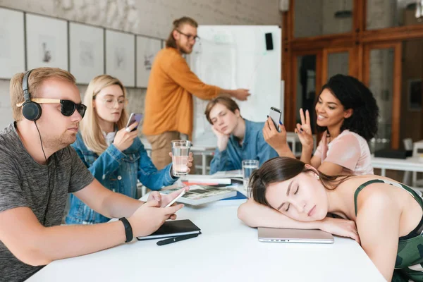 Unga lärare förklara något nära anslagstavla medan studenter som sitter vid bordet och titta inte på honom — Stockfoto