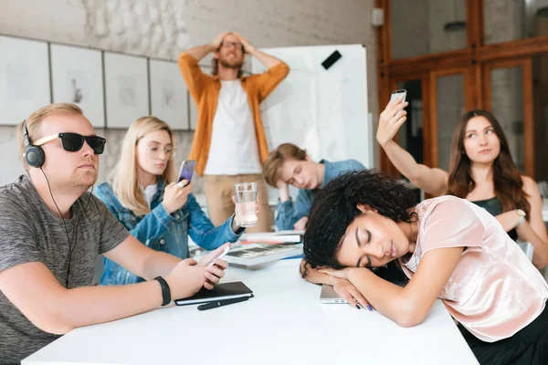 Profesor cansado de pie cerca de la tabla y sosteniendo sus manos en la cabeza. Grupo de jóvenes estudiantes haciendo fotos, escuchando música y durmiendo durante la lección —  Fotos de Stock