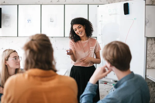 Beautiful African American lady with dark curly hair standing near board and happily looking at her colleagues in office — Stock Photo, Image