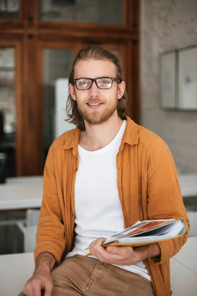 Portrait of nice boy with blond hair and beard in glasses looking in camera while holding journals in hands — Stock Photo, Image