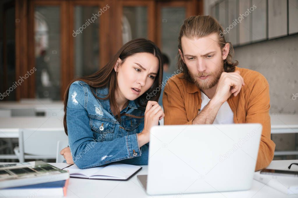 Portrait of young man and girl amazedly looking on laptop. Students sitting at classroom and studying together with laptop and books on table
