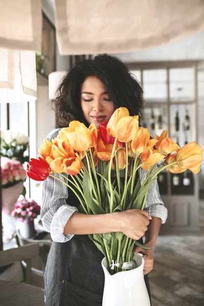 Nice African American girl standing with bouquet of tulips in hands. Portrait of lady with dark curly hair thoughtfully closing her eyes with flowers — Stock Photo, Image