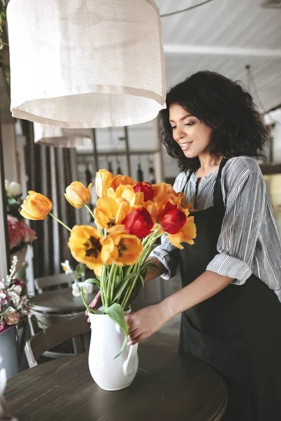 Young African American girl standing near flowers on table. Pretty florist in apron creating bouquet of tulips — Stock Photo, Image