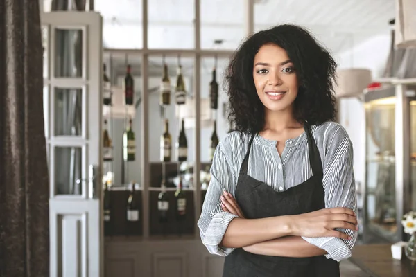 Молодая афроамериканка в фартуке, стоящая со сложенными в ресторане руками. Милая девушка с темными вьющимися волосами, стоящая в фартуке в кафе — стоковое фото