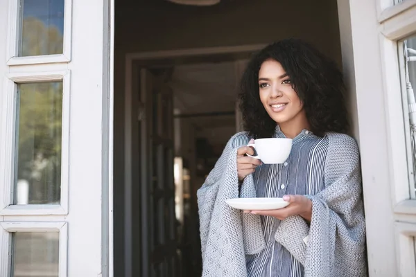 Jovem afro-americana com cabelo encaracolado escuro bebendo café inclinado na porta. Linda senhora de pé com copo na mão envolto em xadrez — Fotografia de Stock
