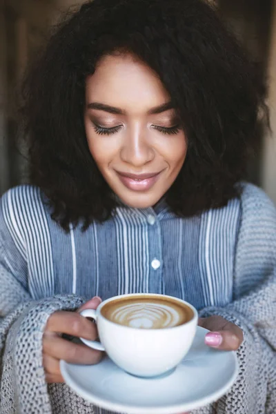 Retrato de bela menina afro-americana com xícara de cappuccino nas mãos. Senhora bonita com cabelo encaracolado escuro cuidadosamente fechando os olhos com xícara de café nas mãos — Fotografia de Stock