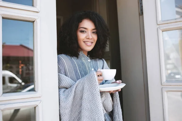 Senyum gadis Afrika-Amerika bersandar di pintu dengan secangkir kopi dibungkus kotak-kotak. Wanita cantik dengan rambut keriting gelap minum kopi di restoran — Stok Foto