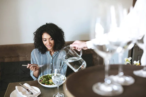 Hermosa chica afroamericana sentada en un restaurante y comiendo ensalada mientras el camarero vierte agua en un vaso — Foto de Stock