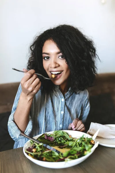 Nice African American girl eating salad in restaurant. Portrait of smiling lady with dark curly hair and salad on table at cafe