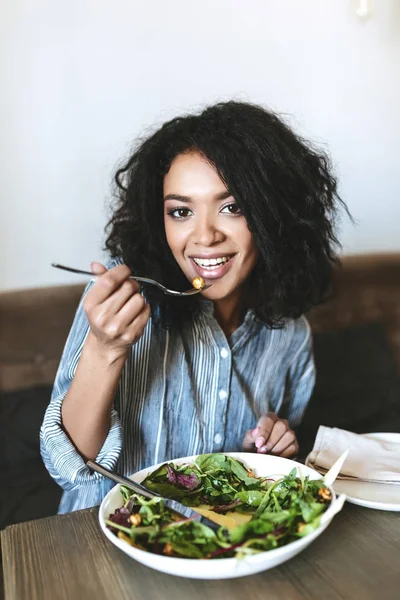 Pretty African American girl eating salad in restaurant. Portrait of smiling lady with dark curly hair and salad on table at cafe