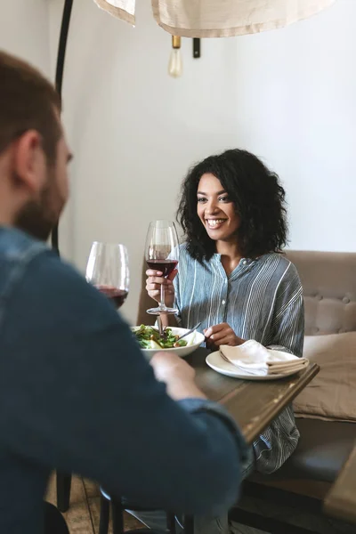 Casal jovem sentado no restaurante e bebendo vinho tinto. Menina afro-americana comendo salada e bebendo vinho no café — Fotografia de Stock