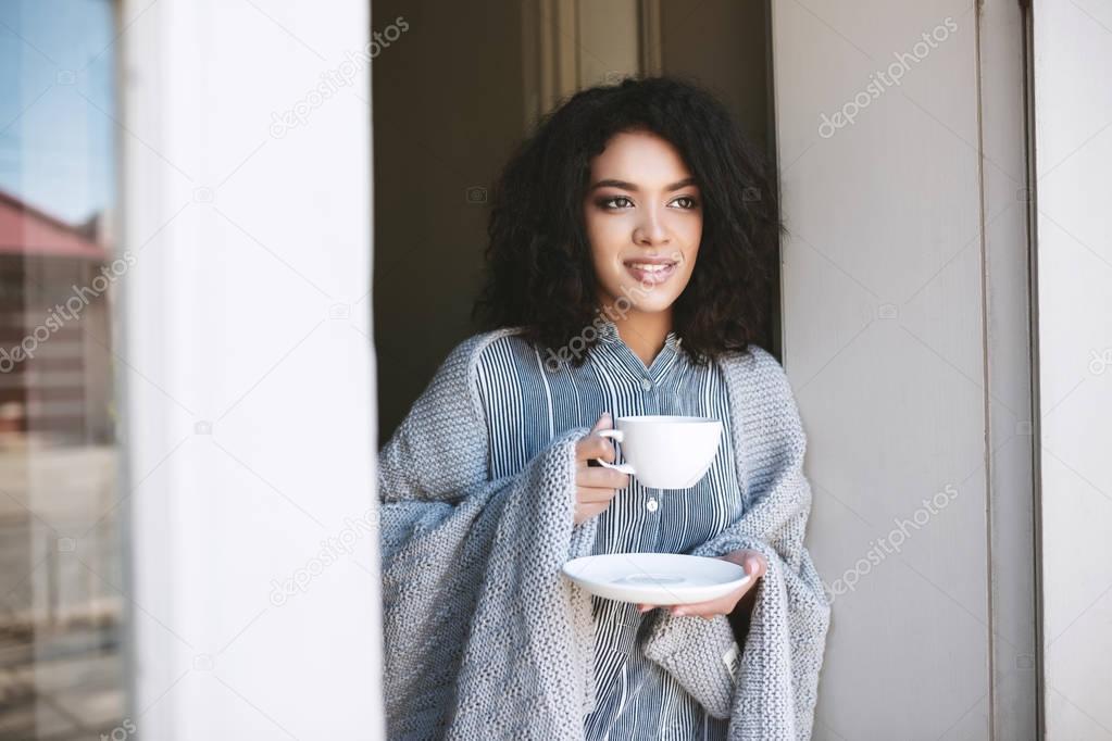 Portrait of beautiful lady with dark curly hair leaning on door with cup of coffee in hands