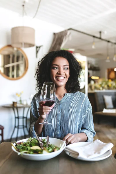 Retrato de chica sonriente con cabello rizado oscuro sentada en restaurante con copa de vino tinto y ensalada en la mesa —  Fotos de Stock