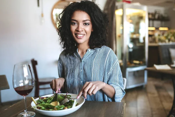 Joven chica afroamericana con el pelo rizado oscuro sentado en el restaurante. Chica sonriente mirando en cámara con copa de vino tinto y ensalada en la mesa en la cafetería —  Fotos de Stock