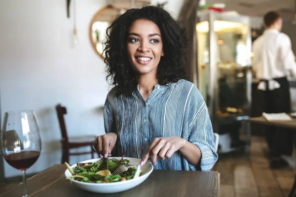 Nice African American girl with dark curly hair eating in restaurant. Smiling girl sitting at cafe and looking aside with glass of red wine and salad on table