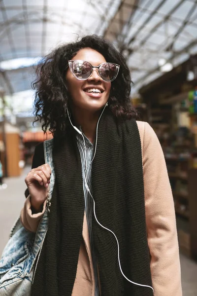 Krásná africká americká dívka stojící na ulici v sluchátka. Portrét cool Girl s tmavými kudrnatými vlasy sluneční brýle chodit po ulicích — Stock fotografie
