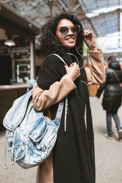 Menina afro-americana sorridente usando óculos de sol e fones de ouvido. Retrato de menina agradável com cabelos encaracolados escuros em pé na rua — Fotografia de Stock