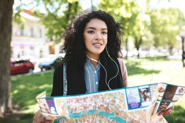 Chica joven en los auriculares cuidadosamente mirando a un lado con el mapa en las manos en la calle — Foto de Stock