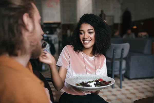 Chica alegre sentada en el restaurante y hablando con un amigo. Sonriente dama afroamericana sentada en la cafetería con plato de ensalada en la mano — Foto de Stock