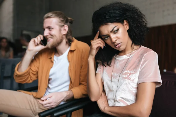 Enojada chica afroamericana con el pelo rizado oscuro tristemente mirando a un lado mientras chico con el pelo rubio felizmente hablando en su teléfono celular en la cafetería —  Fotos de Stock