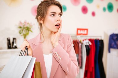 Genç kız pembe ceket amazedly bir kenara alışveriş torbaları el giyim mağazası ile arıyorum. Sürpriz Bayan boutique Satılık giysi raf arka plan ile ayakta