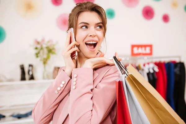 Mosolygó lány rózsaszín portréja kabát, boldogan félre nézett színes bevásárló táskák, váll és mozgatható telefon-ban odaad ruhaüzlet — Stock Fotó