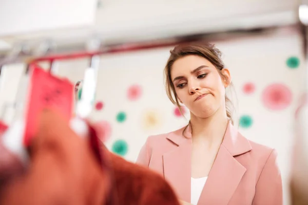 Chica joven en chaqueta rosa y camiseta blanca cuidadosamente mirando en perchero de ropa en boutique —  Fotos de Stock