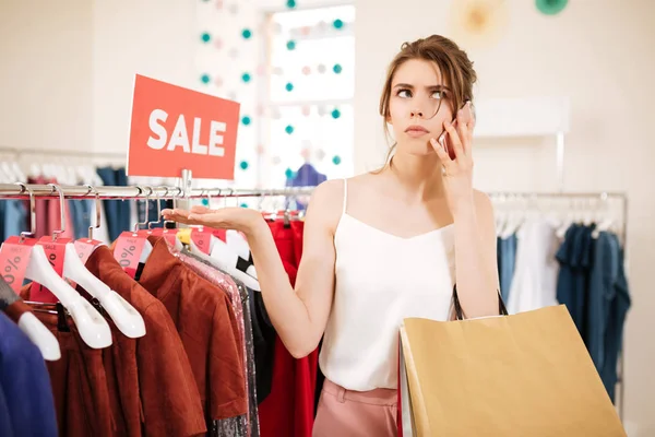 Молодая девушка в белом, стоящая рядом со стойкой для одежды и задумчиво глядя в сторону, разговаривая по мобильному телефону в бутике — стоковое фото