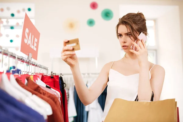 Junges Mädchen in weißem Top, das in der Nähe eines Kleiderständers steht und in der Boutique nachdenklich auf ihre Kreditkarte schaut — Stockfoto