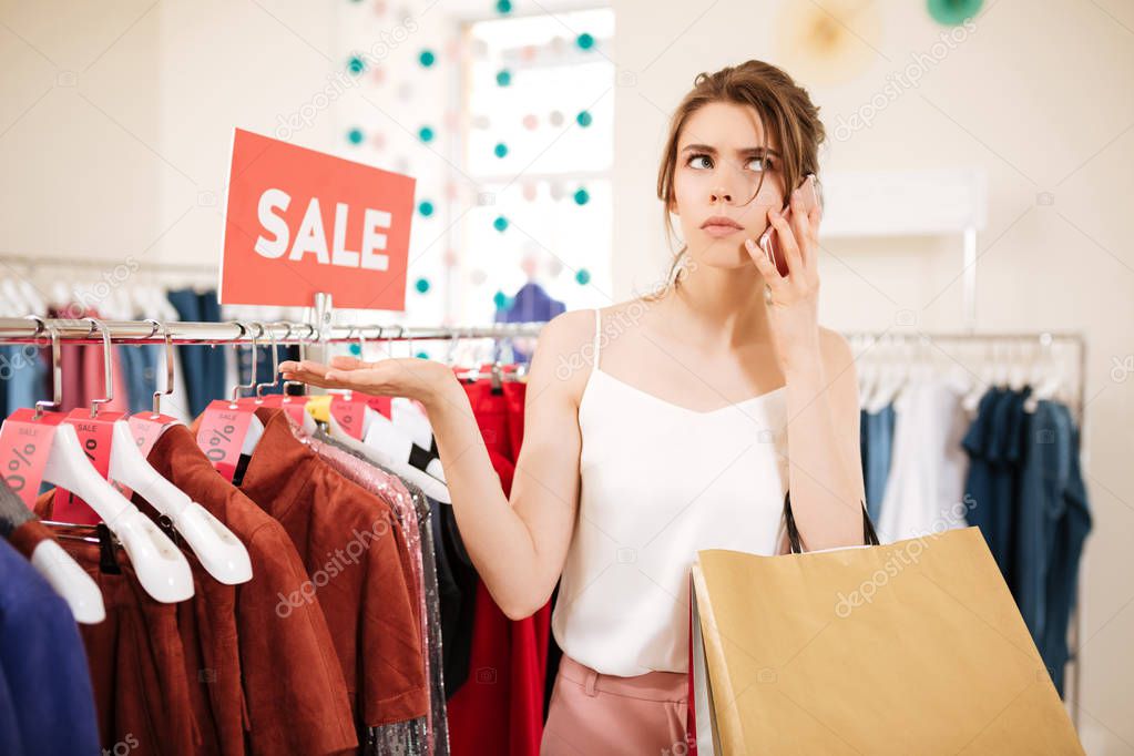Young girl in white top standing near sale clothes rack and thoughtfully looking aside while talking on her cellphone in boutique