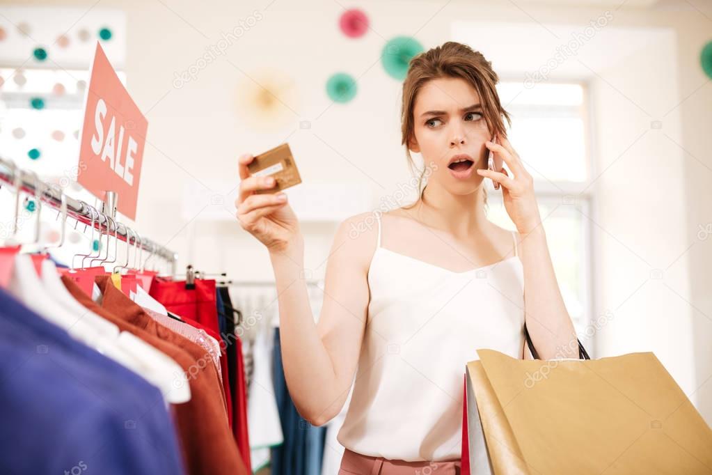 Thoughtful girl in white top standing near sale clothes rack and amazedly looking on her credit card in boutique