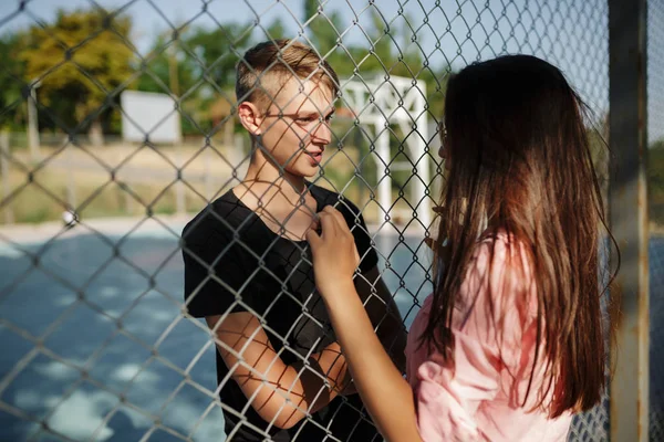 Portrait of young thoughtful boy standing on basketball court and dreamily looking at pretty girl with dark hair through mesh fence — Stock Photo, Image