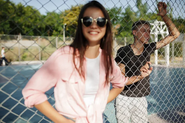 Retrato de chica alegre con pelo oscuro en gafas de sol de pie y mirando en la cámara con valla de malla y niño sonriente en el fondo en la cancha de baloncesto — Foto de Stock