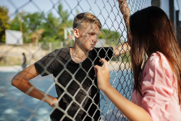 Портрет молодого крутого парня, стоящего на баскетбольной площадке и задумчиво смотрящего на красивую девушку с темными волосами через сетчатый забор — стоковое фото