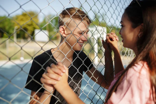 Porträt eines jungen fröhlichen Jungen, der auf dem Basketballplatz steht, während er fröhlich ein nettes Mädchen mit dunklen Haaren durch den Maschendrahtzaun betrachtet und ihre Hand hält — Stockfoto