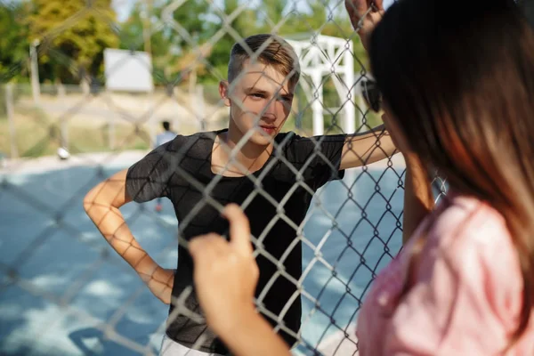 Retrato de menino bonito em pé na quadra de basquete e olhando atenciosamente para a menina com cabelo escuro através de cerca de malha — Fotografia de Stock