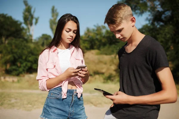Porträtt av ung vacker pojke och flicka som står tillsammans i parken och eftertänksamt letar i mobiltelefoner — Stockfoto
