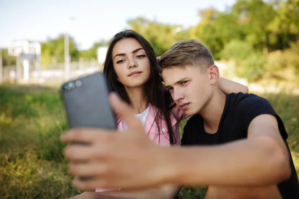 Porträtt av unga vackra paret sitter på gräsmattan i parken och att göra selfie. Söta par att ta foton på frontal mobilkamera — Stockfoto