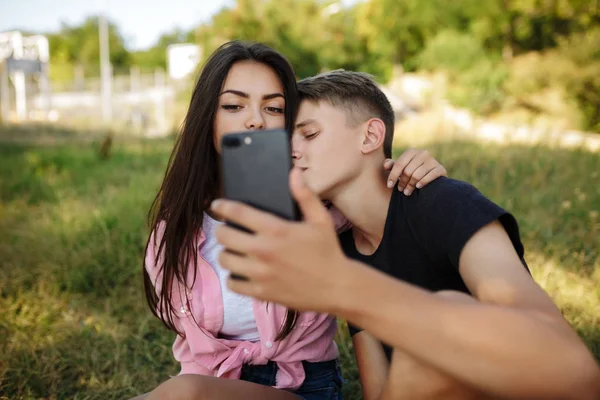 Portrait of young couple sitting on lawn and embracing each other while making selfie in park. Beautiful couple taking photos on cellphone frontal camera — Stock Photo, Image