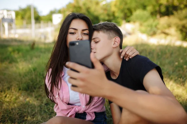 Portrét mladý pár sedí na trávníku a objímat sebe přitom selfie v parku. Krásný pár fotografování na mobil čelní fotoaparát — Stock fotografie