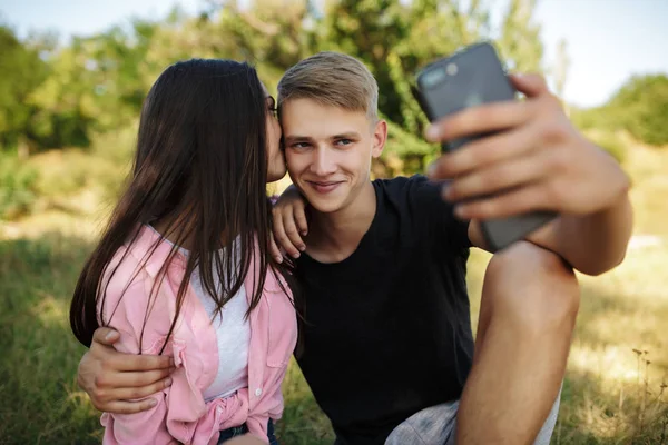 Porträtt av ung cool pojke sitter på gräsmattan i parken och omfamna tjej samtidigt selfie. Söta par att ta foton på frontal mobilkamera — Stockfoto