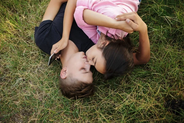 Портрет молодого крутого парня и красивой девушки с сотовым телефоном в руке лежит на зеленой лужайке в парке и целуется — стоковое фото