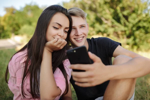 美丽微笑的情侣坐在公园草坪上的肖像, 并愉快地使用手机在一起 — 图库照片
