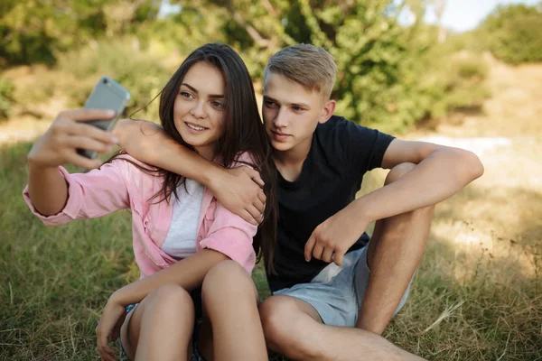 Porträtt av unga vackra paret sitter på gräsmattan i parken och gör selfie tillsammans. Söta par att ta foton på frontal mobilkamera — Stockfoto